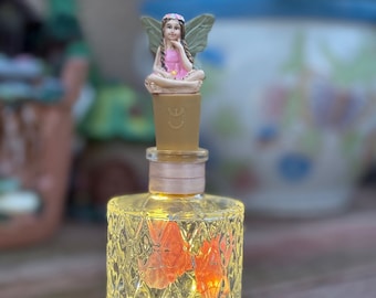 May include: A decorative glass bottle with a fairy bottle stopper. The bottle is illuminated with small lights and features two orange butterfly accents. The fairy figurine is seated with wings and a pink dress. The bottle is on a wooden surface.