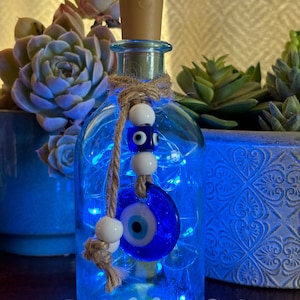 May include: A clear glass bottle with a blue evil eye charm and white beads hanging from a twine string. The bottle is filled with blue fairy lights. The bottle is sitting on a wooden surface.