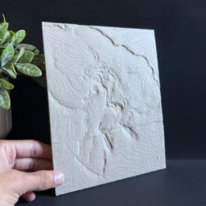 Op de afbeelding: Een beige, rechthoekige reli&euml;fkaart met gedetailleerde topografische lijnen. De kaart heeft een getextureerd oppervlak en wordt in een hoek gehouden, waarbij het driedimensionale ontwerp wordt getoond. De achtergrond is donker en benadrukt de kenmerken van de kaart.