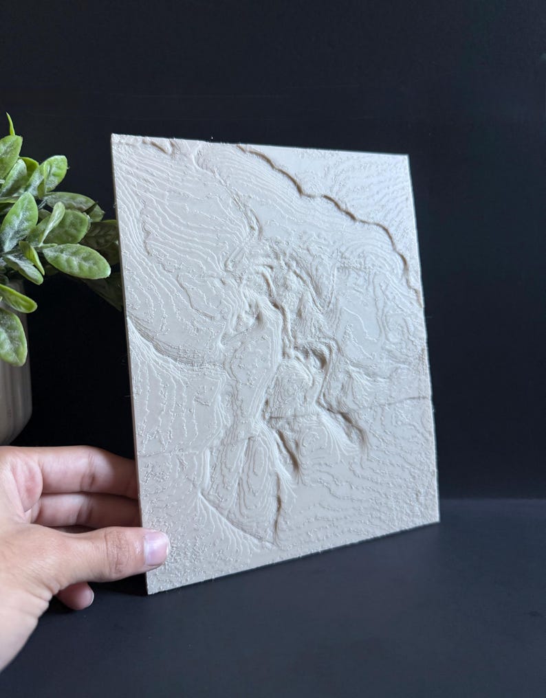 Op de afbeelding: Een beige, rechthoekige reli&euml;fkaart met gedetailleerde topografische lijnen. De kaart heeft een getextureerd oppervlak en wordt in een hoek gehouden, waarbij het driedimensionale ontwerp wordt getoond. De achtergrond is donker en benadrukt de kenmerken van de kaart.