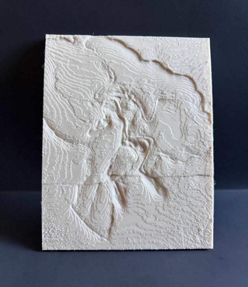 Op de afbeelding: Een rechthoekige, cr&egrave;mekleurige 3D-reli&euml;fkaart met gedetailleerde topografische lijnen. Het verhoogde oppervlak toont een landschap met vari&euml;rende hoogtes, waardoor een textuur ontstaat. De kaart staat tegen een donkere achtergrond.