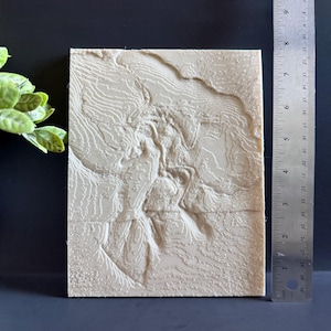 Op de afbeelding: Een beige, rechthoekige 3D topografische kaart met gedetailleerde contourlijnen. De kaart is ongeveer 20 cm hoog en 15 cm breed. Een liniaal is naast de kaart geplaatst voor de schaal. De kaart ligt op een donkere ondergrond.