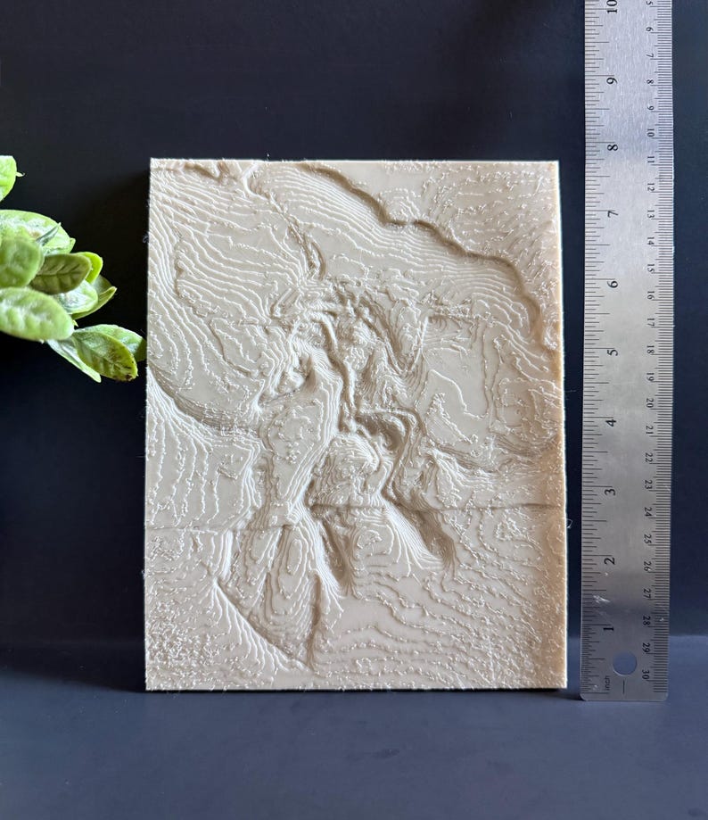 Op de afbeelding: Een beige, rechthoekige 3D topografische kaart met gedetailleerde contourlijnen. De kaart is ongeveer 20 cm hoog en 15 cm breed. Een liniaal is naast de kaart geplaatst voor de schaal. De kaart ligt op een donkere ondergrond.