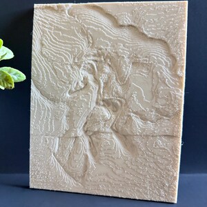 Op de afbeelding: Een beige, rechthoekige reli&euml;fkaart met gedetailleerde topografische lijnen. Het 3D-geprinte kunstwerk toont een landschap met vari&euml;rende hoogtes, waardoor een textuur ontstaat. Het stuk wordt gepresenteerd tegen een donkere achtergrond, wat het ingewikkelde ontwerp benadrukt.