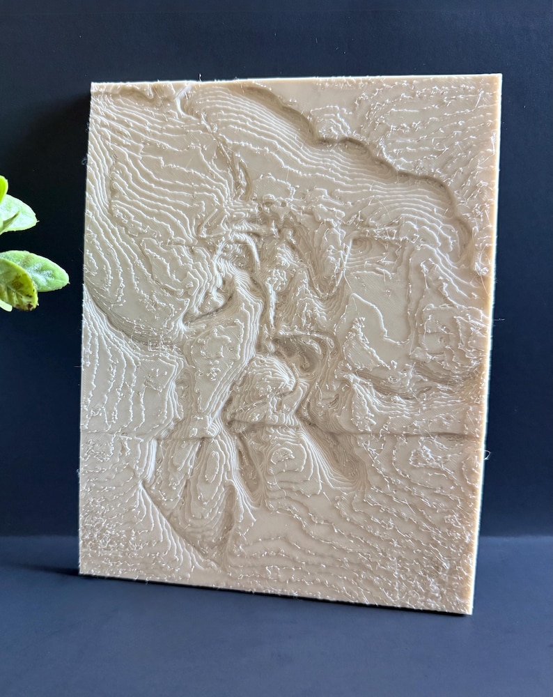 Op de afbeelding: Een beige, rechthoekige reli&euml;fkaart met gedetailleerde topografische lijnen. Het 3D-geprinte kunstwerk toont een landschap met vari&euml;rende hoogtes, waardoor een textuur ontstaat. Het stuk wordt gepresenteerd tegen een donkere achtergrond, wat het ingewikkelde ontwerp benadrukt.