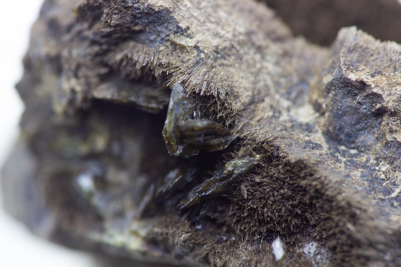 May include: Close-up of a mineral specimen with a rough, textured surface. The rock is primarily dark gray and brown, with crystalline structures and a fibrous texture. The mineral has a natural, earthy appearance.