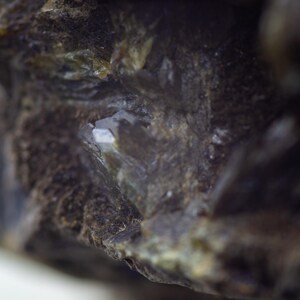May include: Close-up of a rough, dark brown mineral with a crystalline structure. The mineral has a textured surface with areas of green and white, and a small, reflective, crystal-like formation.