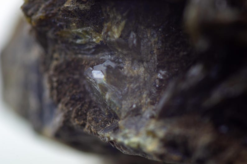 May include: Close-up of a rough, dark brown mineral with a crystalline structure. The mineral has a textured surface with areas of green and white, and a small, reflective, crystal-like formation.
