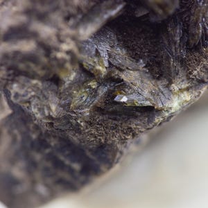 May include: Close-up of a rough, dark brown rock formation with crystalline structures. The rock has a textured surface with areas of green and small, reflective crystal formations. The background is a blurred white.