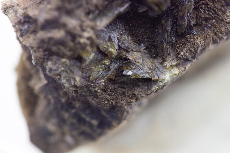 May include: Close-up of a rough, dark brown rock formation with crystalline structures. The rock has a textured surface with areas of green and small, reflective crystal formations. The background is a blurred white.