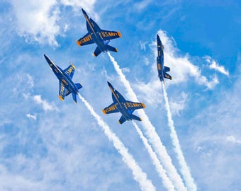 May include: Four blue and yellow U.S. Navy fighter jets flying in a formation against a bright blue sky with scattered white clouds. Each aircraft features the text U.S. NAVY on the wings and leaves a long, distinct white smoke trail behind it.
