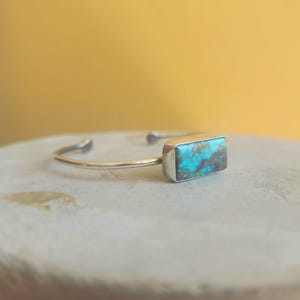 May include: A silver cuff bracelet featuring a rectangular turquoise stone with brown matrix patterns. The bracelet has a smooth, polished metal band that is open at the back, showcasing a vibrant blue-green gemstone set in a simple metal frame.