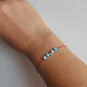 Può includere: Un braccialetto di corda rossa con perline quadrate di colore turchese e un piccolo ciondolo dorato.