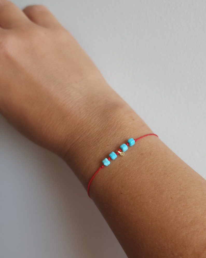K&ouml;nnte beinhalten: Ein d&uuml;nnes rotes Armband aus Kordel mit vier kleinen, leuchtend t&uuml;rkisfarbenen w&uuml;rfelf&ouml;rmigen Perlen und einem winzigen, runden goldfarbenen Anh&auml;nger, der zwischen den Perlen platziert ist und ein zartes Accessoire bildet.