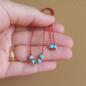 K&ouml;nnte beinhalten: Ein rotes Kordelarmband mit vier kleinen t&uuml;rkisfarbenen w&uuml;rfelf&ouml;rmigen Perlen und einem winzigen goldfarbenen runden Anh&auml;nger. Das Armband enth&auml;lt zwei weitere t&uuml;rkisfarbene w&uuml;rfelf&ouml;rmige Perlen an den Enden der verstellbaren Kordel.