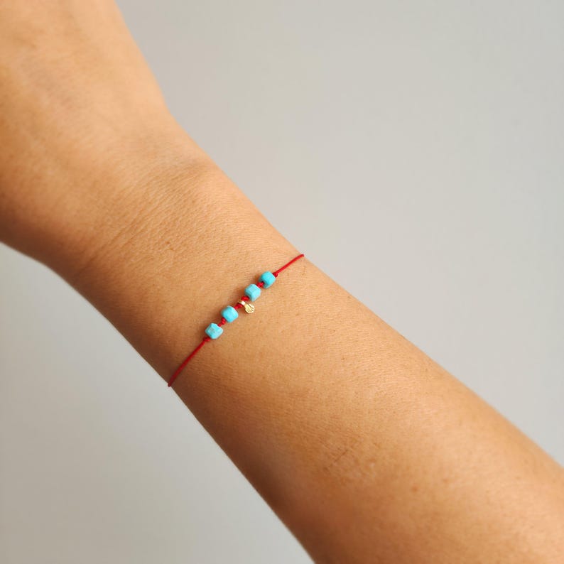K&ouml;nnte beinhalten: Ein zartes Armband aus rotem Faden mit vier kleinen, quadratischen, leuchtend t&uuml;rkisfarbenen Perlen und einem winzigen, runden, goldfarbenen Anh&auml;nger, der mittig am Band befestigt ist. Das Armband liegt um ein Handgelenk und zeigt ein schlichtes Design.