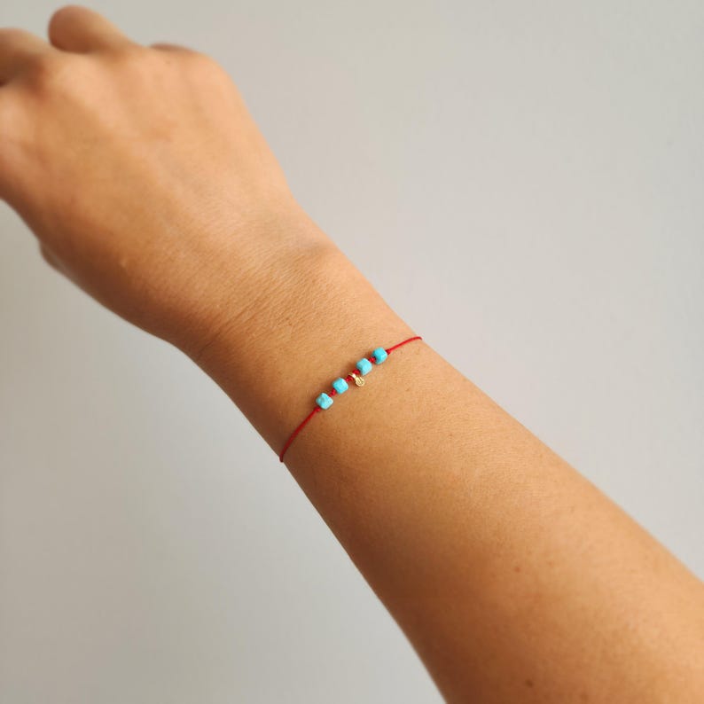 K&ouml;nnte beinhalten: Ein zartes Armband aus roter Schnur mit vier kleinen, quadratischen, leuchtend t&uuml;rkisfarbenen Perlen und einem winzigen, runden, goldfarbenen Metallanh&auml;nger, die mittig auf dem d&uuml;nnen Band angeordnet sind und ein dezentes Accessoire bilden.