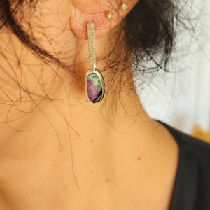 May include: Silver-tone drop earrings with a rectangular textured top and a bezel-set, oval-shaped stone. The stone features a mix of colors, including green and pink, creating a unique and eye-catching design. These earrings are a stylish accessory.