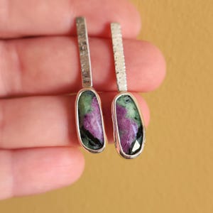 Könnte beinhalten: Silberne Ohrringe mit strukturiertem Stab und einem ovalen Stein. Der Stein hat grüne, violette und schwarze Farbtöne. Die Ohrringe werden vor einem gelben Hintergrund gezeigt.