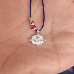 May include: A silver necklace with a small, detailed pendant featuring a smiling face. The necklace is on a dark blue cord with white and red beads.