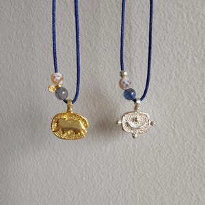 Könnte beinhalten: Zwei Halsketten mit Anhängern an blauen Schnüren. Der Anhänger links ist golden und der Anhänger rechts ist silbern. Beide Anhänger haben eine strukturierte Oberfläche.