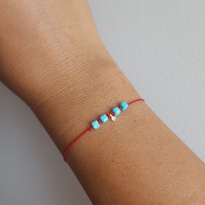 K&ouml;nnte beinhalten: Ein d&uuml;nnes rotes Kordelarmband mit vier kleinen, quadratischen, leuchtend t&uuml;rkisfarbenen Perlen und einem winzigen, runden, goldfarbenen Metallanh&auml;nger, der zwischen den Perlen angebracht ist. Das Armband ist mit einem einfachen Knoten versehen.