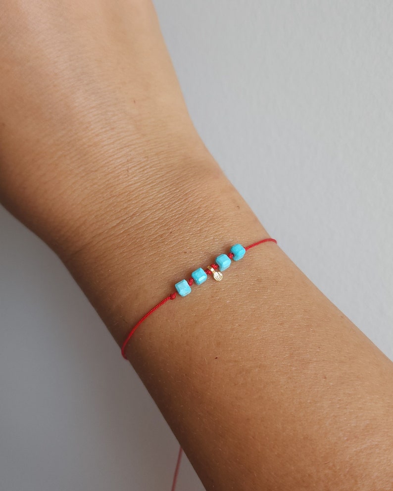 K&ouml;nnte beinhalten: Ein d&uuml;nnes rotes Kordelarmband mit vier kleinen, quadratischen, leuchtend t&uuml;rkisfarbenen Perlen und einem winzigen, runden, goldfarbenen Metallanh&auml;nger, der zwischen den Perlen angebracht ist. Das Armband ist mit einem einfachen Knoten versehen.