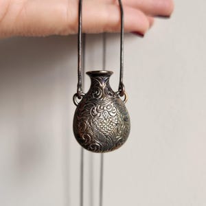 Puede incluir: Un pequeño collar con colgante plateado con un diseño de botella de perfume grabado y detallado. La botella tiene forma bulbosa con cuello estrecho y dos asas pequeñas. El intrincado patrón incluye motivos florales y de búho.