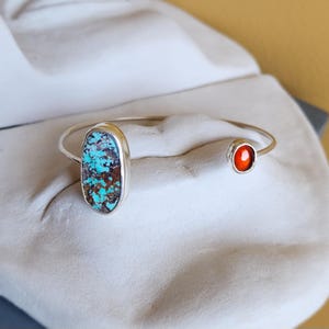 Könnte beinhalten: Ein silbernes Armband in offenem Design mit zwei verschiedenen Steinen. Ein Ende ist mit einem großen, ovalen Türkis mit dunkelbraunen Matrixmustern besetzt, während das andere Ende einen kleineren, runden, leuchtend orange-roten Stein trägt.