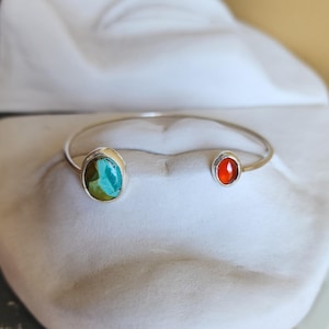 May include: A silver open cuff bracelet featuring two distinct stones. One end holds a larger, oval-shaped turquoise stone with blue and green marbling, while the other end features a smaller, round, vibrant orange carnelian stone.