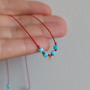 K&ouml;nnte beinhalten: Eine rote Kordelkette mit vier kleinen, quadratischen, leuchtend t&uuml;rkisfarbenen Perlen und einem kleinen, strukturierten, goldfarbenen runden Anh&auml;nger, der mittig zwischen den Perlen sitzt. Die Kordelenden sind mit zwei weiteren t&uuml;rkisen Perlen versehen.