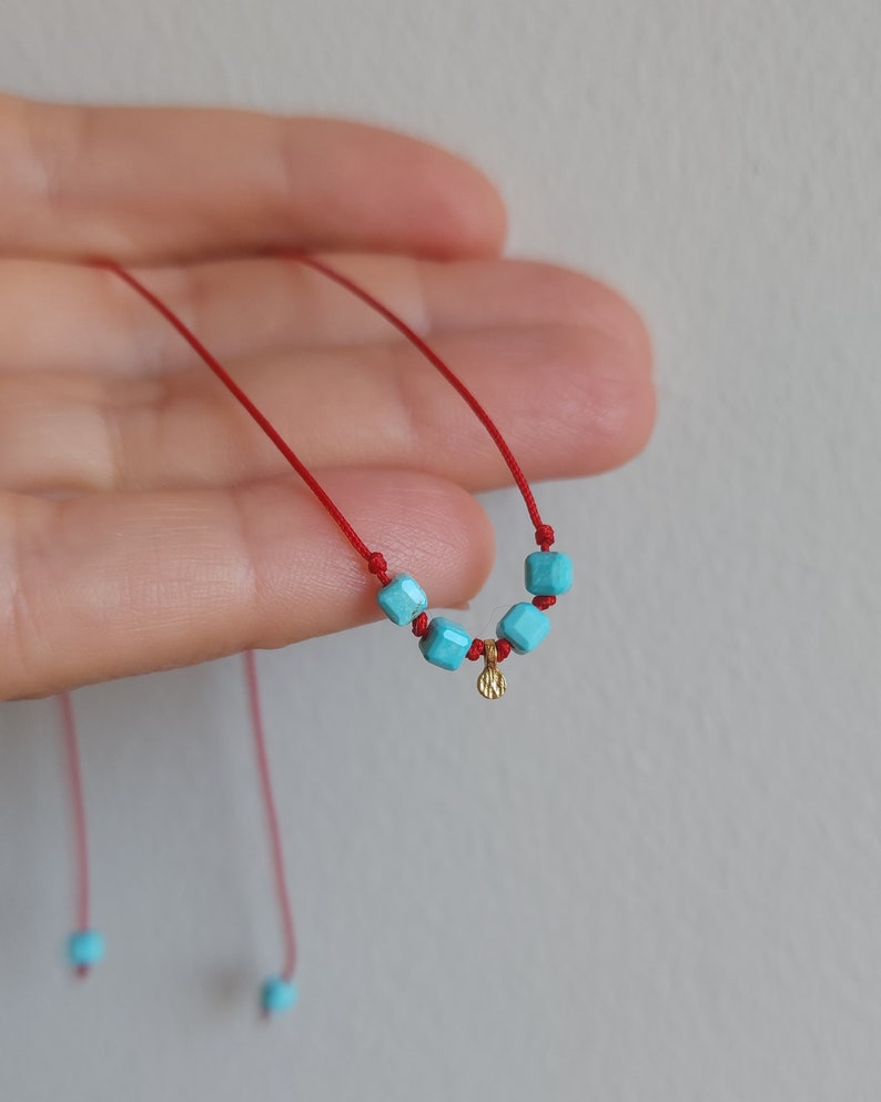 K&ouml;nnte beinhalten: Eine rote Kordelkette mit vier kleinen, quadratischen, leuchtend t&uuml;rkisfarbenen Perlen und einem kleinen, strukturierten, goldfarbenen runden Anh&auml;nger, der mittig zwischen den Perlen sitzt. Die Kordelenden sind mit zwei weiteren t&uuml;rkisen Perlen versehen.