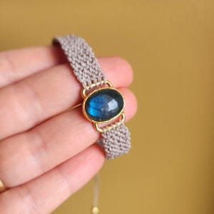 May include: A bracelet featuring a central oval-shaped labradorite gemstone with a blue iridescent sheen, set in a gold-toned metal frame. The band is constructed from light beige woven cord, connecting to the central stone setting on both sides.