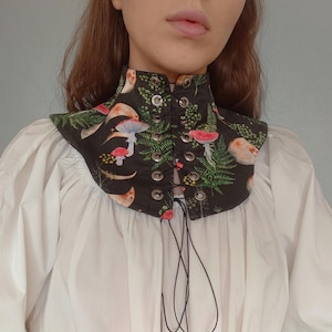 Può includere: Un colletto nero con lacci con stampa di funghi e felci. Il colletto è in tessuto e ha una chiusura con lacci neri sul davanti.