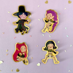 Op de afbeelding: Vier goudkleurige stickers met stripfiguren in verschillende poses. Elk personage heeft een unieke outfit en kapsel. De stickers staan op een lichtpaarse achtergrond met hart- en halvemaanvormige confetti.