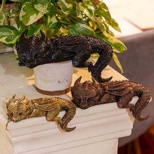 Könnte beinhalten: Drei Drachenfiguren in Schwarz, Bronze und Gold. Der schwarze Drache ruht auf einem weißen Topf, die anderen liegen auf einer weißen Oberfläche. Jeder Drache hat detaillierte Flügel und Hörner.