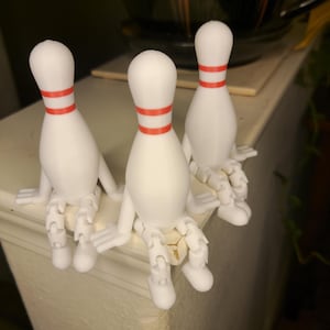 May include: Three white bowling pin figurines with red stripes sitting on a white surface. Each figurine has articulated arms and legs.