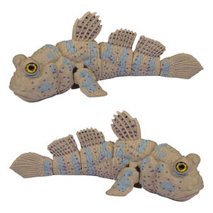 May include: Two articulated mudskipper fish figures featuring a beige base color with light blue patches and small purple spots. Each figure has prominent yellow eyes with black pupils, textured skin, and segmented bodies that allow for movement.