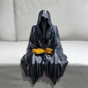 May include: A black 3D printed figurine of a hooded figure sitting with a book and a cup in their lap.