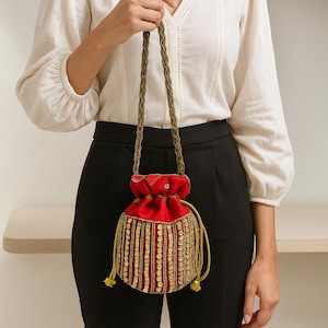 May include: A red drawstring bag with gold embellishments and a braided strap. The bag features a vibrant red fabric with intricate gold sequin and bead detailing. The drawstring closure is accented with gold tassels.