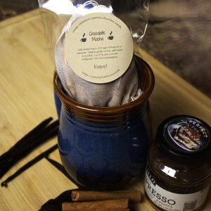 May include: A brown paper bag with a white muslin tea bag inside. The bag is labeled "Chocolate Mocha" and includes instructions for brewing the tea. The bag is sitting in a blue and brown ceramic mug. There are also cinnamon sticks, a chocolate bar, and a jar of espresso powder.