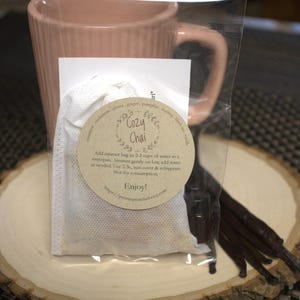 Puede incluir: Una bolsa de plástico transparente que contiene una bolsa de muselina blanca con una etiqueta marrón que dice "Cozy Chai" e incluye instrucciones de uso. La bolsa está junto a una pequeña botella de vidrio de líquido y varias vainas de vainilla sobre una superficie de madera.