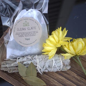 May include: A wooden tray containing a white pouch labelled "Clean Slate" with a tea bag, a bundle of white sage smudge sticks, and two bay leaves. Yellow flowers are in the background.
