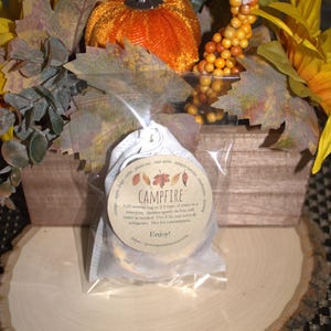 May include: A small white pouch with a brown label that reads "CAMPFIRE" and instructions for use. The pouch is sitting on a wooden slice with fall-themed decorations.