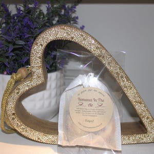May include: A gold heart-shaped frame with a white satin bag inside. The bag contains a small round tag with the text "Romance In The Air" and a heart symbol. The tag also says "Enjoy!"