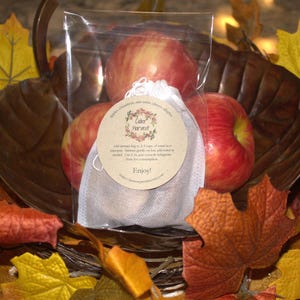 Może przedstawiać: Biały woreczek z muślinu wypełniony czerwonymi jabłkami, z brązową papierową etykietą z napisem "Cider Harvest" i "Enjoy!" Woreczek znajduje się w brązowym koszyku z jesiennymi liśćmi.