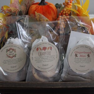 Puede incluir: Tres pequeñas bolsas de muselina blanca con etiquetas para los aromas "Color Harvest", "Campfire" y "Gingerbread Man". Las bolsas están colocadas en una bandeja de madera con follaje otoñal y una calabaza.