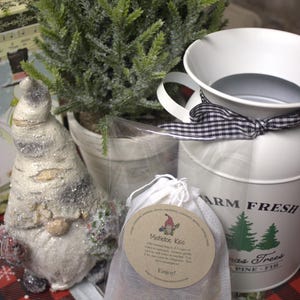 May include: A white bag with a brown label that reads "Mistletoe Kiss" and instructions for use. The bag is sitting on a table with a red and white checkered tablecloth. There is a small white gnome figurine and a white pitcher with a black and white checkered ribbon in the background.