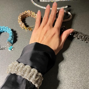 May include: Various sparkling jewellery items, including bracelets and hair accessories, are arranged on a dark surface. A silver beaded bracelet is on a black sleeve. Other bracelets are gold, silver, and turquoise. Silver hair accessories are also present.