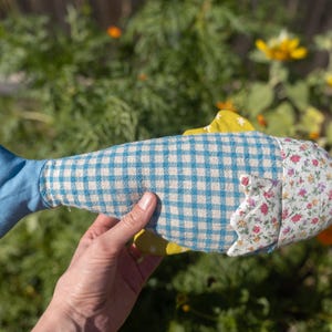 May include: A stuffed fish toy featuring a blue tail, a central body section with a light blue and white gingham pattern, and a head section covered in a white fabric with a small floral print. The toy includes yellow fabric fins.